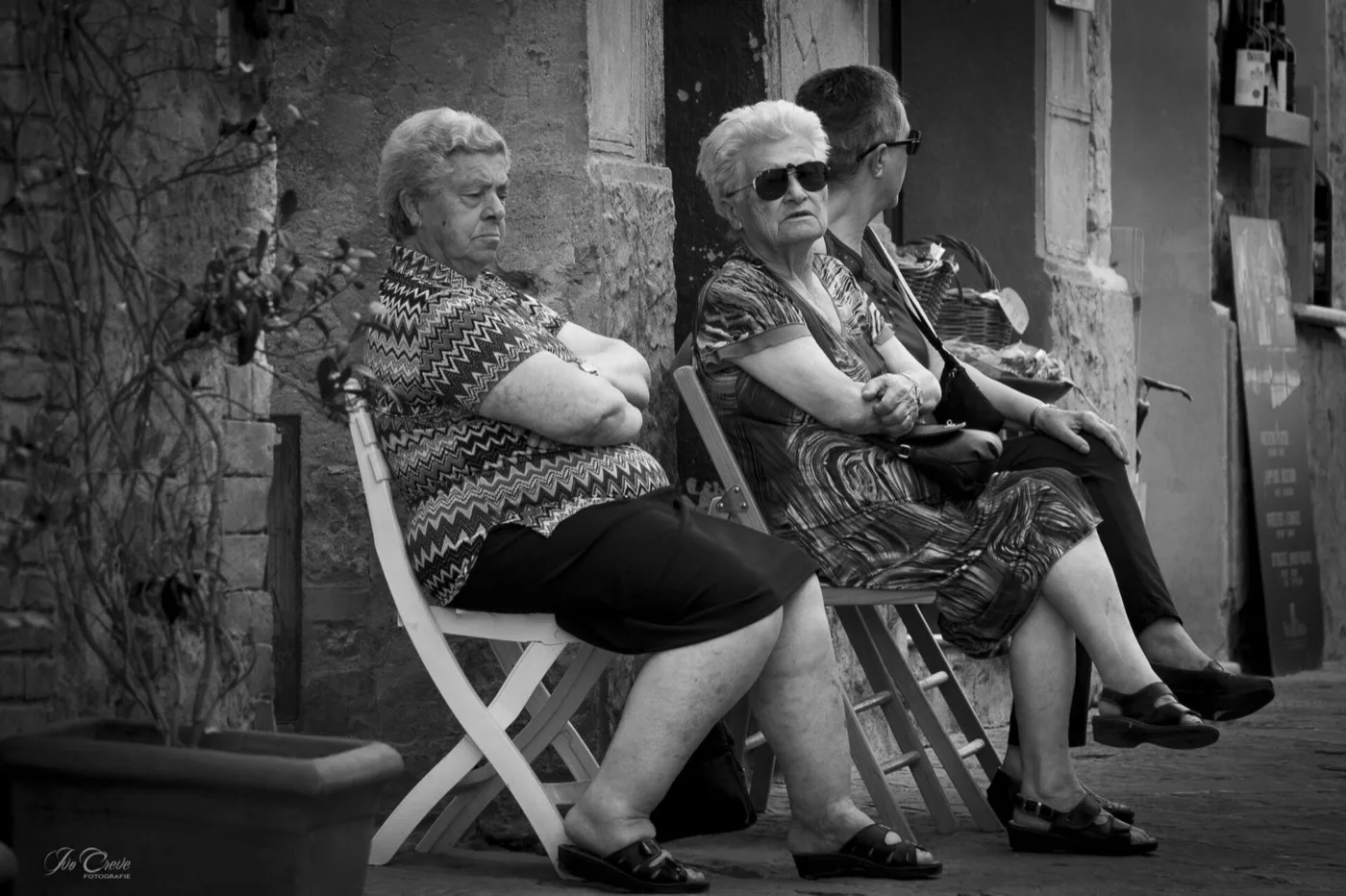Drie ouderen op stoelen voor een gevel, in zwart-wit